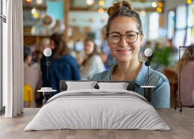 A young woman smiles confidently in a vibrant cafe setting, showcasing casual attire and glasses while surrounded by people. Wall mural