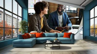 African businessman in eyeglasses smiling to his colleague during conversation while they working over project at table Wall mural