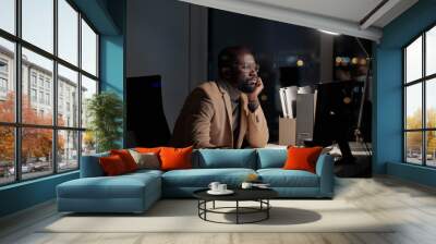 African businessman sirring by desk in front of computer monitor in office while working late at night Wall mural