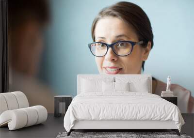 Attractive businesswoman in stylish glasses talking to colleague while sitting near light blue wall.  Wall mural