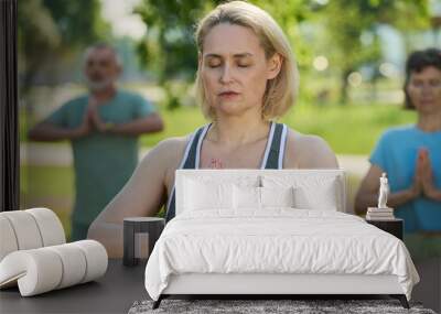 Blond female yoga instructor keeping eyes closed while practicing exercises against elderly people repeating after her Wall mural