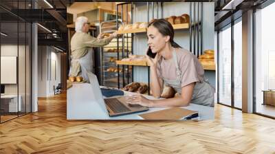 Busy female in apron using laptop and talking to clients on the phone by workplace Wall mural