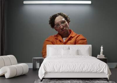 Captured image of a man wearing glasses and an orange jumpsuit holding handcuffs in an indoor setting Wall mural