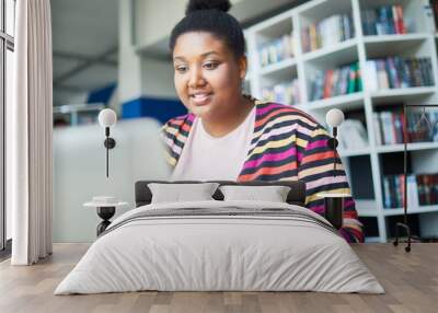 Cheerful confident young African-American overweight student reading online article on laptop while preparing for exam in library Wall mural