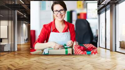 Cheerful young woman with toothy smile looking at you while tying ribbon on giftbox Wall mural