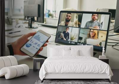 Close-up of businessman working in empty office and using smartphone to view graph while discussing sales with colleagues via video conferencing app Wall mural