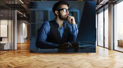 Concentrated entrepreneur or manager sitting by desk in front of computer monitor while working at night Wall mural