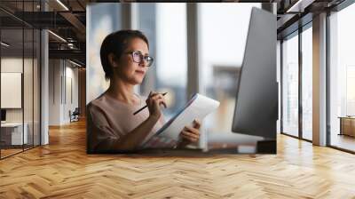 confident businesswoman with document looking at computer screen and making working notes in office Wall mural