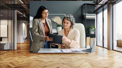 Confident mature female boss looking through document held by young secretary and checking points while sitting by her workplace Wall mural