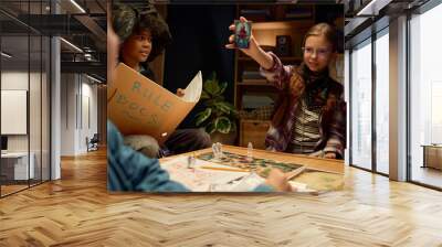 Cute girl in eyeglasses and casual attire showing card with octopus or other character to friends while playing tabletop game Wall mural