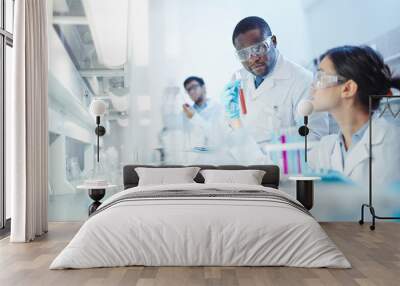 Female Asian laboratory scientist in lab coat and safety goggles showing test tube with red liquid to curious African-American colleague in laboratory. Latin-American scientist in background. Wall mural