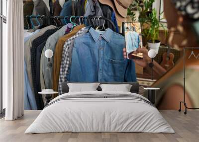 Focus on secondhand denim shirt hanging in front of row of casual attire on rack while young female consumer taking picture of apparel Wall mural