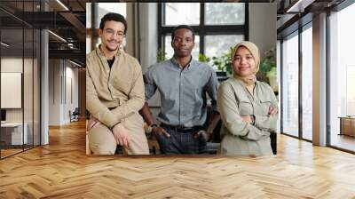 Group of three young multicultural employees in casual attire standing in row in front of camera in openspace office and looking at you Wall mural