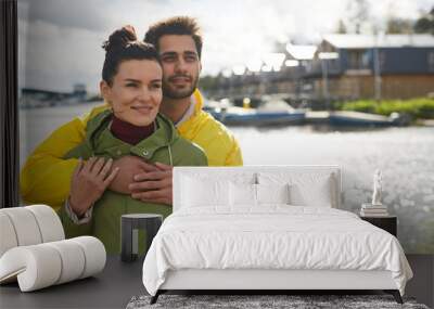 Happy dreamy young couple in coats standing together at seaport and looking at view while contemplating seascape together, smiling man hugging girlfriend Wall mural