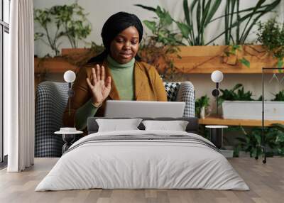 Happy young black woman with laptop on her knees sitting in armchair and waving hand to patient during online psychological session Wall mural