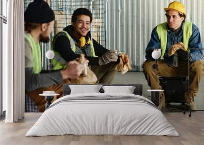 Happy young engineer with sandwich and cup of hot tea chatting to colleagues at lunch break while sitting in front of them in warehouse Wall mural