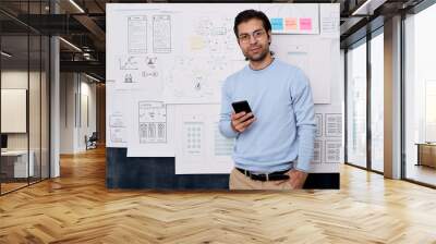 Horizontal medium portrait shot of young Caucasian man working in IT industry standing with smartphone looking at camera Wall mural