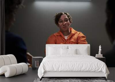 Inmate in orange jumpsuit sitting at a table with hands extended, participating in an interrogation in a dimly lit room with two unseen individuals interacting Wall mural