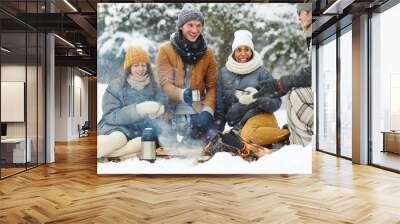 Joyful young people in winterwear sitting in snow arounf fire and having hot drinks Wall mural