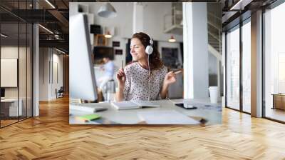 Joyous young businesswoman with headphones listening to dynamic music while working by desk in front of computer monitor Wall mural