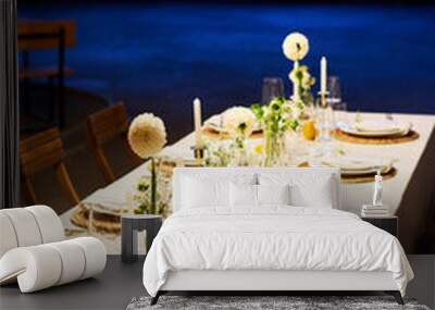 Long table covered with white tablecloth with dahlia flowers, candles, decorations and wineglasses surrounded by two rows of plates Wall mural