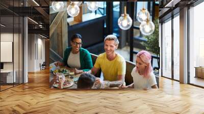 One of guys sitting by served table between two intercultural girls during lunch and talking to one of friends Wall mural