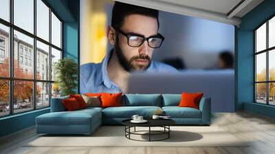 Portrait of handsome bearded man wearing glasses working with laptop in dark office late at night, his face lit up by screen Wall mural