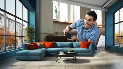 Portrait of handsome emotional adult man playing video game holding wireless controller and  smiling joyfully while sitting on couch at home Wall mural