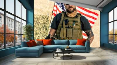 Portrait of serious brutal bearded man in vest with radio device holding riffle against American flag outdoors Wall mural