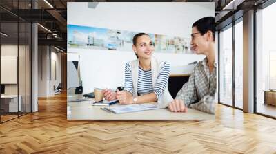 Portrait of two businesswomen, one mature and one young, sitting at desk together in modern office, discussing something and smiling during meeting Wall mural