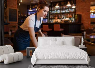 Pretty young waitress in apron using detergent and duster to clean table Wall mural