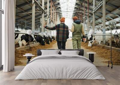 Rear view of two young workers of modern cowfarm walking along cowsheds and discussing conditions of livestock maintain Wall mural