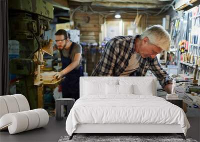 Senior master of woodworking processing detail with chisel while his young trainee working by machine Wall mural