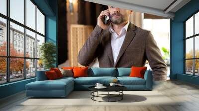 Serious agent with mobile phone listening to his client while standing by table Wall mural