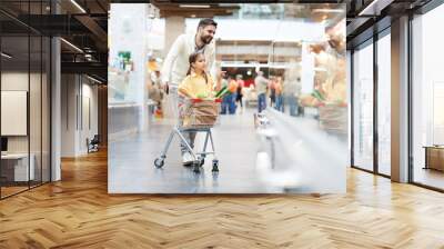 Serious girl sitting in shopping cart and pointing with finger at shelf while helping father with shopping in supermarket Wall mural