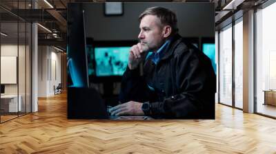 Serious male security guard concentrating on watching CCTV video on screen of computer while sitting by workplace in observation room Wall mural