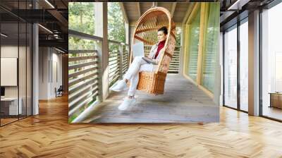 Serious pensive young lady freelancer in white sneakers sitting in hanging wicker chair and using laptop while working on balcony Wall mural