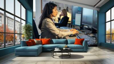 Side view of young Hispanic female operator of surveillance system sitting by desk in front of computer monitors in office Wall mural