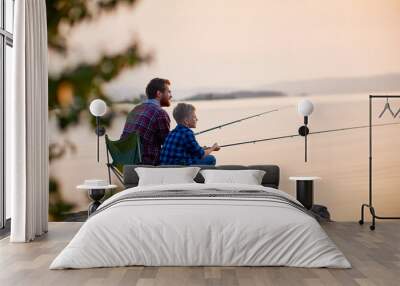 Side view portrait of father and son sitting together on rocks fishing with rods in calm lake waters with landscape of setting sun, both wearing checkered shirts, shot from behind tree Wall mural