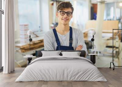 Smiling confident handsome young joiner in uniform standing in modern workshop and crossing arms on chest while looking at camera Wall mural