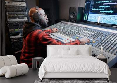 Stylish African American music producer wearing headphones sitting at mixing desk recording song in studio Wall mural