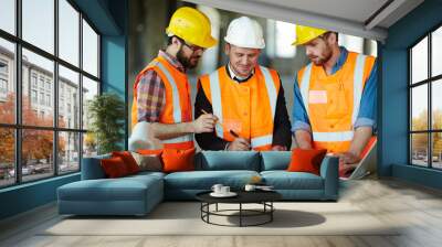 Team of construction workers wearing protective helmets and vests discussing project details with executive supervisor standing at table with blueprints Wall mural