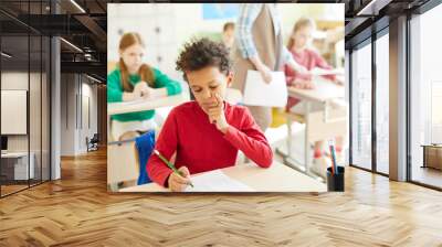 Thoughtful elementary student boy with curly hair sitting at desk and passing test in classroom, he leaning on hand Wall mural