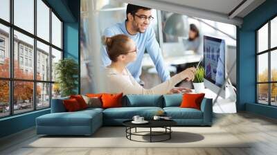 Two colleagues having business discussion in modern office, woman analyst showing successful financial and marketing statistics from screen to her male co-worker, sitting at desk with laptop computer Wall mural