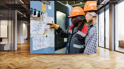 Two factory workers examining detailed blueprints pinned on a corkboard discussing important aspects of the technical drawings in an industrial setting Wall mural