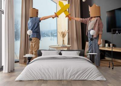 Two little siblings in casualwear and cardboard helmets playing with toy swords Wall mural