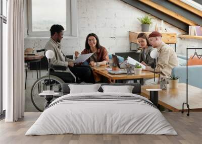 Two pairs of employees working in small groups by workplace while female economist pointing at document held by male colleague in wheelchair Wall mural