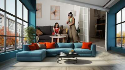 Two women spending leisure time in modern living room, with one seated on sofa and other standing, holding a book. Abstract art adorns walls, creating cozy atmosphere Wall mural