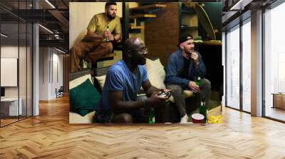 Two young men with beer looking at tv screen and watching video game while black guy with joystick sitting near by and pressing buttons Wall mural