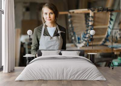 Waist-up portrait of confident fair-haired woodworker with tape measure in hands posing for photography while standing on background of workshop Wall mural